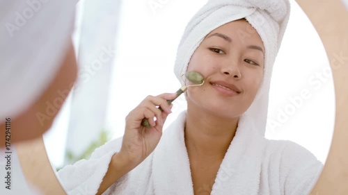 A young Asian woman practices her skin care routine at home, using a jade roller while smiling. She is wrapped in a bathrobe and has a towel on her head, creating a relaxing atmosphere.