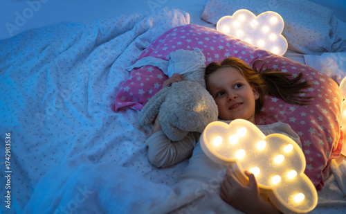 Good night sleep for toddler concept, cute girl with teddy bear and cloud that glow looking at ceiling, copy space
