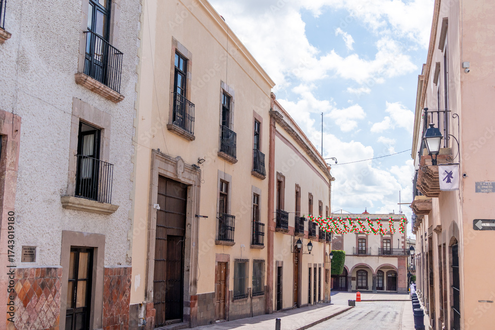 Obraz premium A street with a few buildings and a few people walking around. The street is narrow. Historic center of Querétaro, colonial architecture, decorations for the celebration of Mexico's Independence Day.