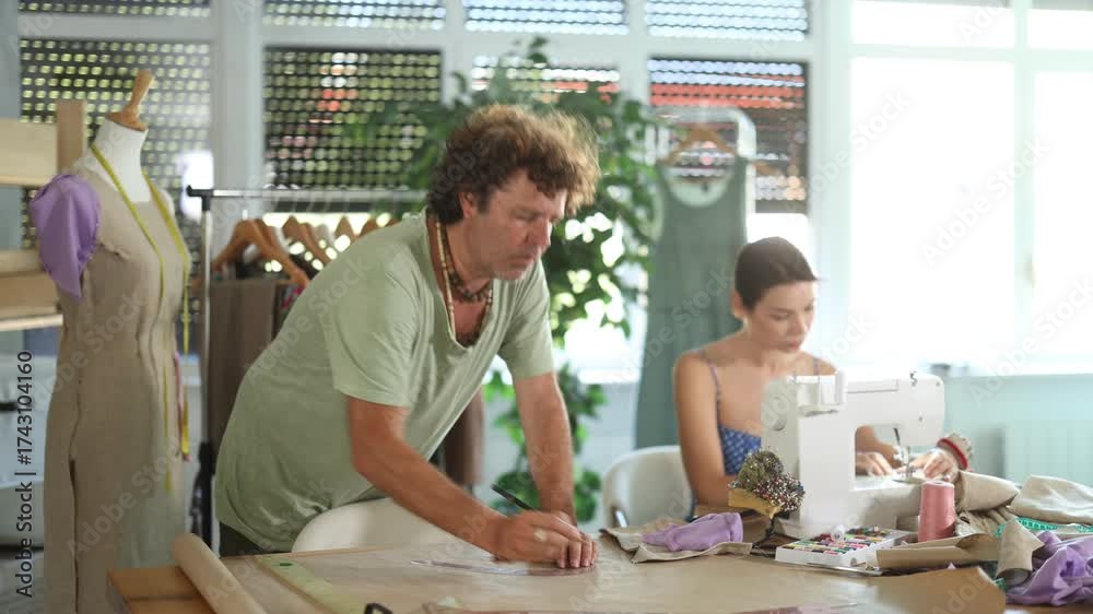 Man tailor draw dress pattern of product on paper using curve ruler, female apprentice seamstress sewing cloth with sewing machine. Family manufactory, piece production of clothing