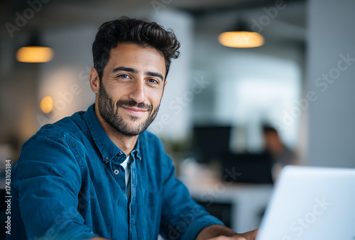 Smiling young man in blue shirt working on laptop in modern office with soft lighting