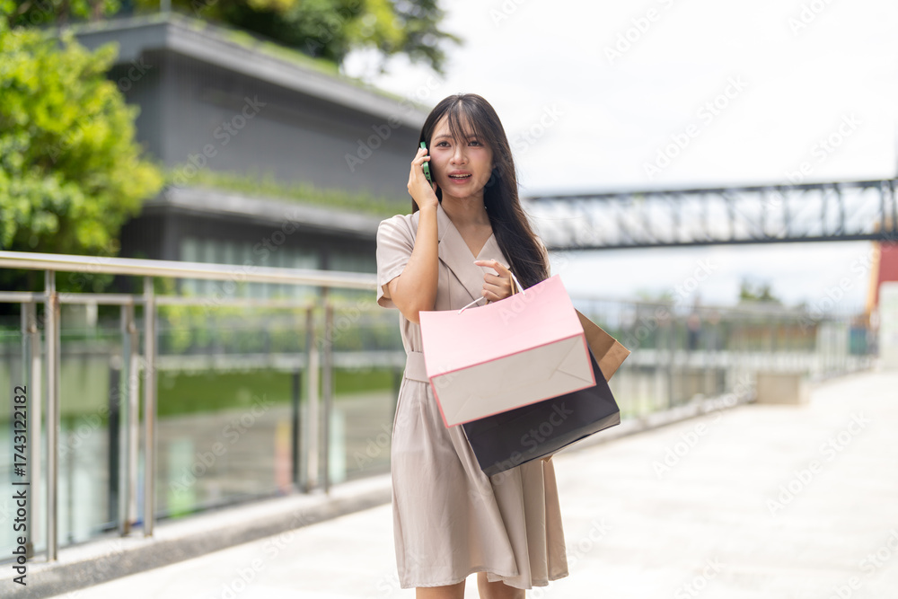 Fototapeta premium Asian woman talk on smartphone holding shopping bags at mall, modern lifestyle fashion shopping, mobile communication, digital age and consumer confidence, trendy online shopping and smart payment