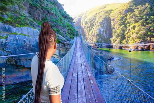 A beautiful young South African girl with braided hair watches the bridge - The suspension bridge over the storms river mouth within Tsitsikamma National Park - Eastern Cape in South Africa