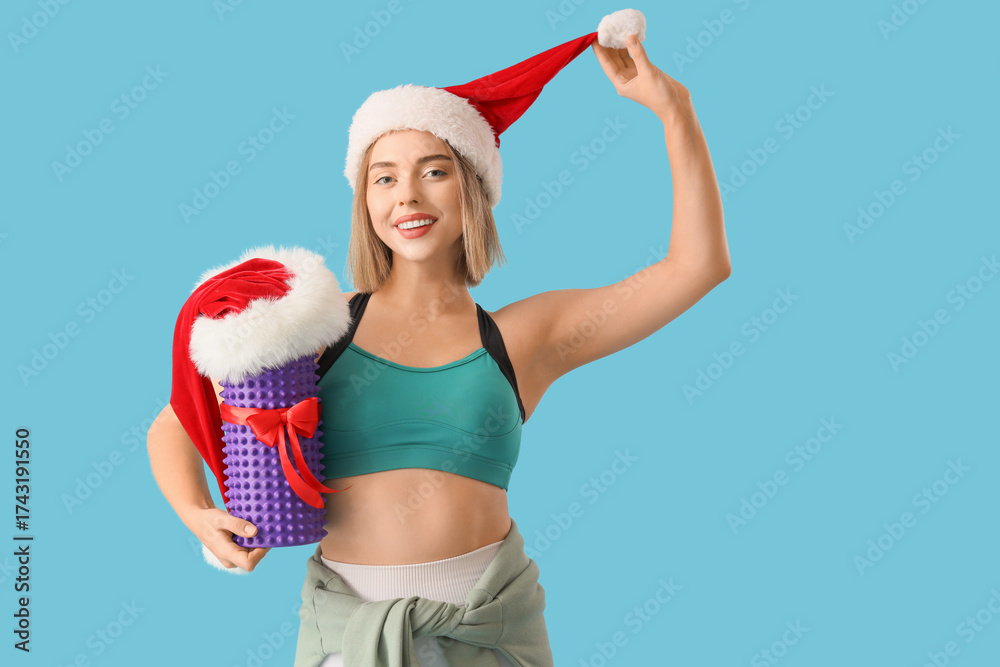 Obraz premium Sporty young woman in Santa hat with body roller as Christmas gift on blue background