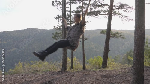 A beautiful young woman in a brown plaid shirt is swinging on a swing in an autumn forest against a backdrop of mountains and hills.