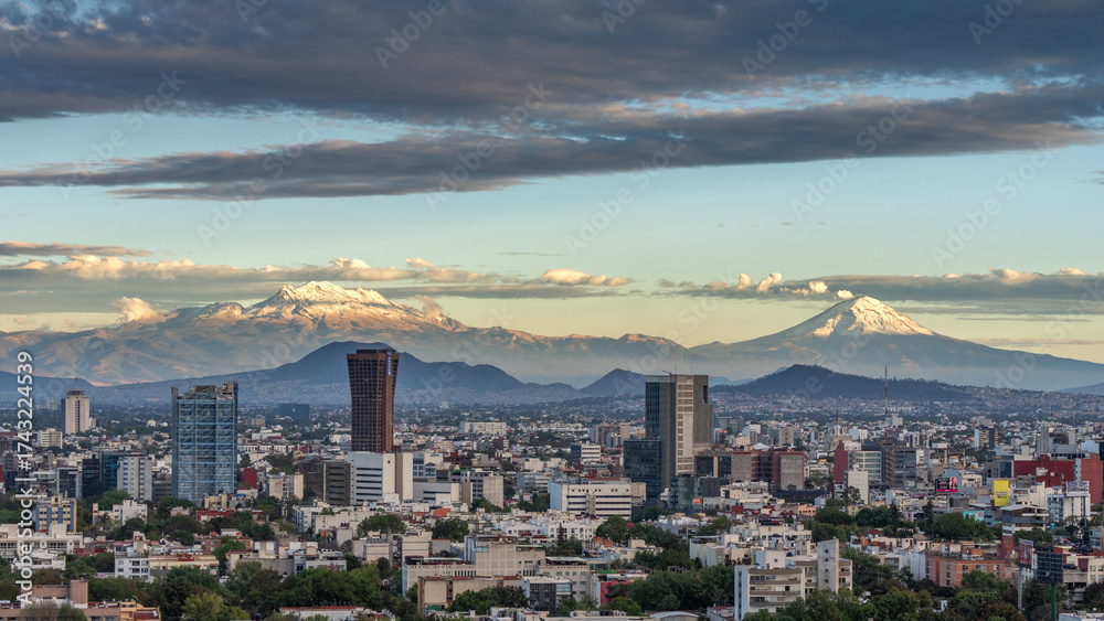 Obraz premium A city with a mountain range in the background. The sky is cloudy and the sun is setting. Panoramic view of Mexico City, snow-covered volcanoes Popocatepetl and Iztaccihuatl