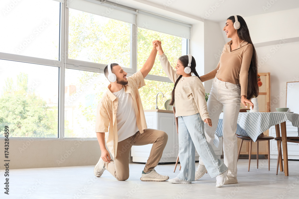 Naklejka premium Happy parents and their little daughter with headphones dancing in kitchen
