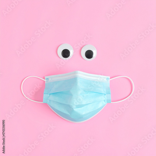 Blue surgical mask with googly eyes arranged in minimal surreal face design on pastel pink background, evoking playful and cheerful healthcare concept