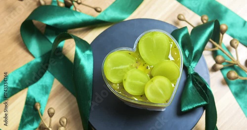Layered heart shaped dessert box presenting kiwi slices, garnished with fresh green grapes and emerald ribbons against dark background