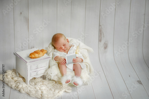 A newborn photo shoot. The baby is 10 days old. The baby is wearing a white robe and fluffy slippers, sitting in a chair. The newborn boy is holding a laptop and a bottle of milk. The younger son 