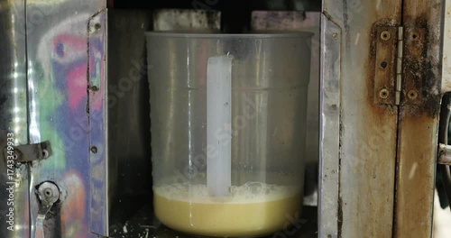 Transparent plastic container with bubbling yellow liquid inside rusty graffitied metal box, revealing improvised scientific or experimental environment