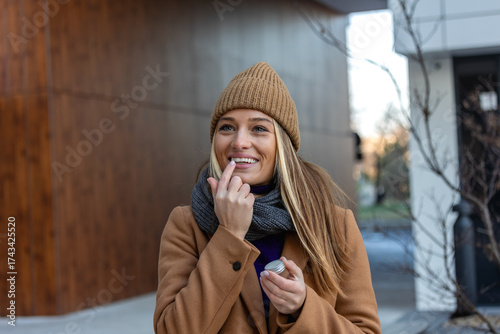 Smiling young woman applying lip balm while outdoors on a cold day. Concept of winter skincare, hydration, beauty routine, and natural wellness.