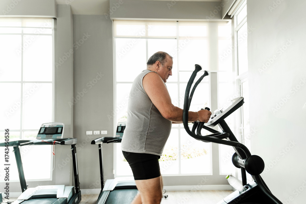 Fototapeta premium Senior man trains on elliptical in bright gym with large windows, sweating through cardio workout. Scene shows strength, motivation, discipline, rehabilitation, recovery, effort, and overall wellness.