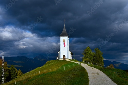 Church of St. Primož and Felician at Jamnik in Gorenjska, Slovenia lit by sunlight and dark clouds in the sky