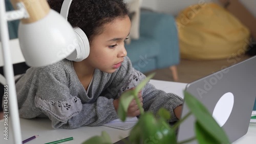 Primary school African American Student child with headphones hand writing in book using laptop. Distance learning online education. School girl studying at home with digital tablet computer and doing