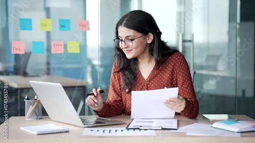 Happy young businesswoman satisfied with financial results looks at computer and document, sitting at workplace in office. Smiling female financier rejoices at positive indicators, celebrates success