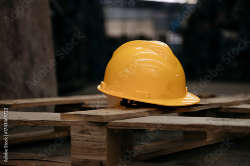 Tablou pe pânză Bright yellow safety hard hat resting on a stack of wooden pallets in a dimly li