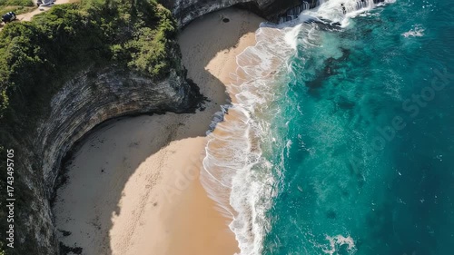 Aerial view of a sandy beach nestled between a rock cliff and turquoise water. Waves roll ashore