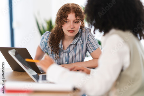 Two women collaborate on a project, one using a laptop while the other points at the screen with a pencil, demonstrating teamwork and shared focus.