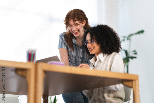 Two women collaborate at a desk, one laughing while looking at a laptop, the other smiling as she works.