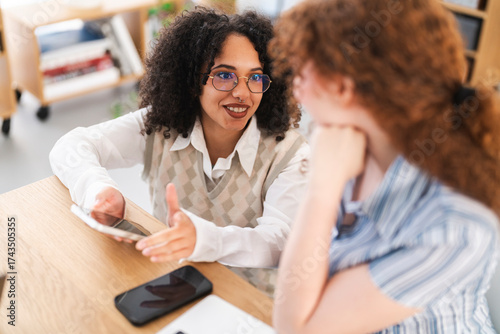 Two young women collaborate at a desk, one holding a tablet and gesturing while the other listens attentively.
