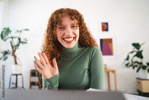 A young woman with curly red hair smiles and waves hello during a video call, working from her home office.