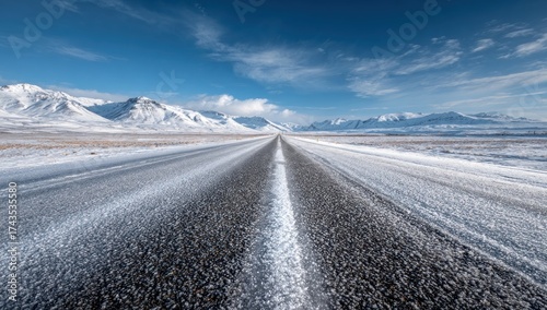 A snowy road stretches into the distance, flanked by snow-covered fields and mountains under a blue sky