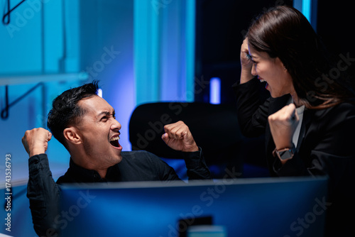 Excited adult male and female tech professionals celebrate night work success with coding AI stock market gains in a dynamic office