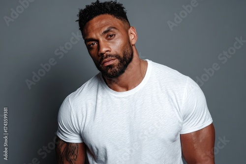 A well-built man with dark skin and a beard gazes directly at the viewer. Background is plain gray