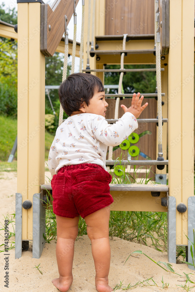 Obraz premium First steps of a cute mixed-race, Latina, and Asian girl with dark hair playing happily in a playground, trying to climb the stairs of a swing while waving her hand.