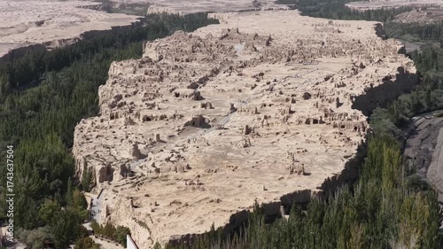 Aerial view of the ancient city ruins with tan structures contrasting against the dark green trees and river, Jiaohe Ancient Town Ruins, Turpan, China.