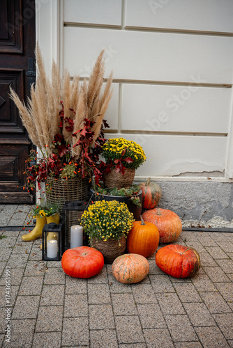 Seasonal autumn pumpkins, potted yellow chrysanthemums, pampas grass, and burning candles creating a cozy fall display by a rustic house entrance on a brick patio