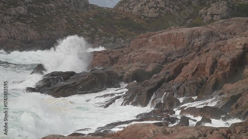 Powerful ocean waves crash against rugged rocks along a wild coastline. The scene captures the raw beauty of nature during stormy weather, creating dramatic splashes and mist.