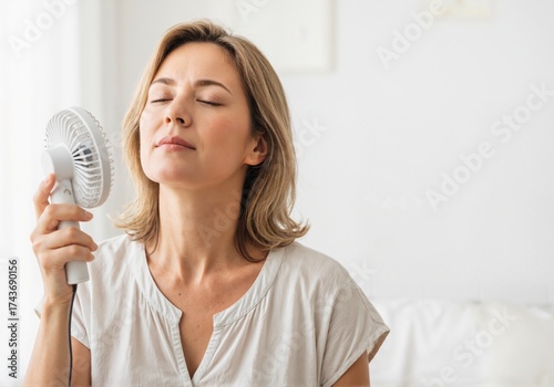 A woman in a light blouse cools herself with a portable fan in a bright room