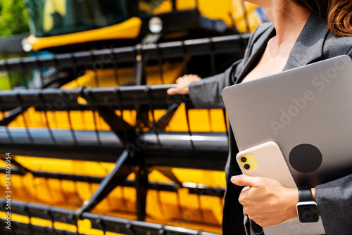 Modern businesswoman holding electronic devices next to combine harvester at dealership.