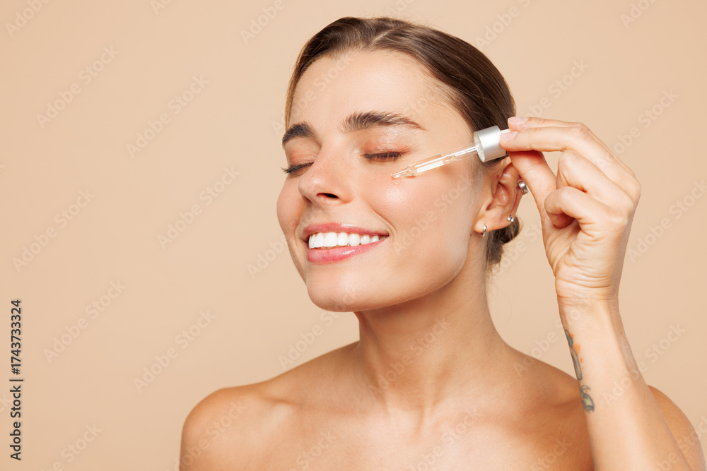 Fototapeta premium Close up beautiful half naked topless young woman 20s with nude make up applying face body oil, moisturizing serum isolated on plain beige background. Skin care healthcare cosmetic procedures concept.