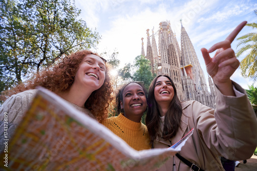 Three cheerful multiracial female tourists holding a map and pointing at something next to the sagrada familia, enjoying holidays together. Vertical image with copy space.