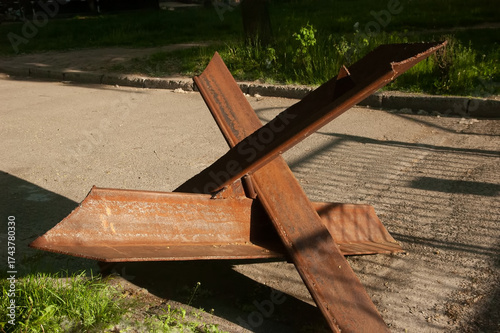 Antitank hedgehog on an asphalt path, sunlight casting shadows from wrought-iron gates. Grass on the sides, a dirt trail branching off, wartime scene.