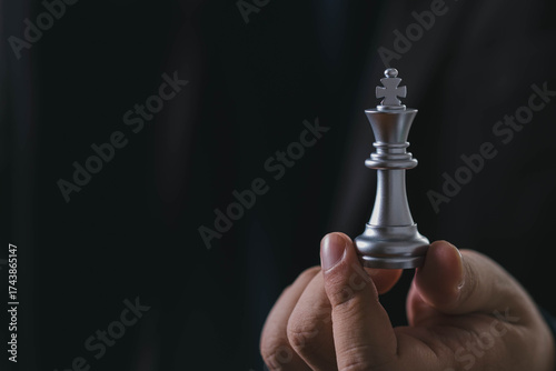 Close-up of a businessman holding silver chess king piece on dark background, symbolizing leadership, strategic decision making, business power, executive control, and authority in management.