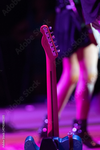 This close-up shot focuses on the neck and headstock of an electric guitar, standing upright on a stage. The guitar's neck is bathed in vibrant pink and purple stage lighting