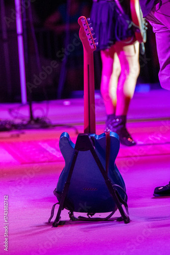 A blue electric guitar stands on a black guitar stand on a stage, illuminated by vivid pink and purple stage lights.