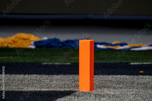 This close-up shot features a bright orange pylon marking a boundary on an artificial turf football field. The pylon stands prominently in the foreground on a striped section of the field