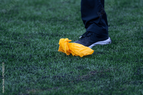 This close-up, low-angle shot captures a person's foot standing on an artificial green turf field, positioned right next to a crumpled yellow penalty flag. The foot is clad in a dark athletic shoe