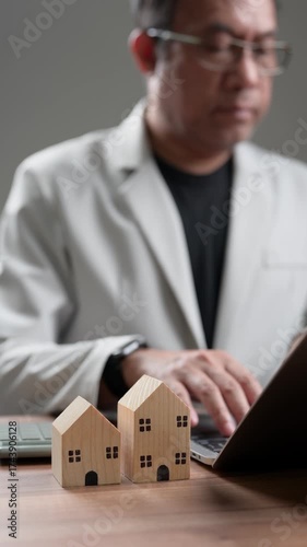 Focused professional analyzes real estate investments, managing personal finances. Small wooden houses represent property ownership, future home planning, wealth growth. Signifies diligent work