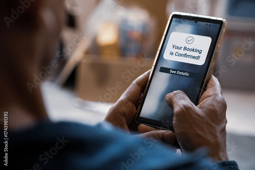 Man using smartphone displaying Your Booking is Confirmed notification on display. Flight or hotel or online appointment booking confirmation message. Selective focus.