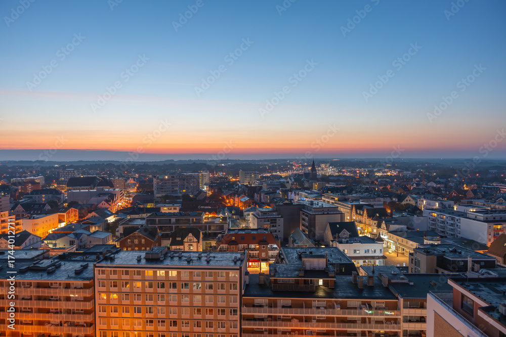 Naklejka premium The town lights glow as night falls, with the sunset fading behind the horizon. Streets and buildings illuminate under the twilight sky.