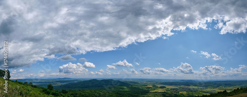 A panoramic view of rolling hills and distant valleys under a dramatic sky with thick clouds and patches of sunlight, revealing a vibrant summer landscape stretching far into the horizon.