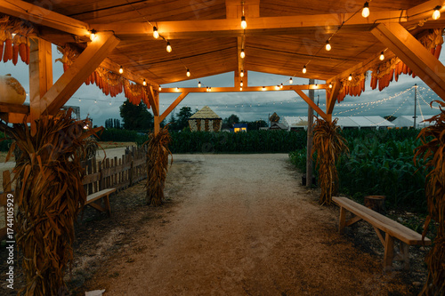 Rustic wooden gazebo lit with warm lights and a bench beneath, set by a countryside road, decorated for Halloween.
