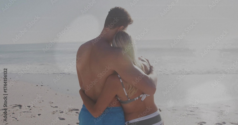 Fototapeta premium Hugging couple in striped bikini and blue swim trunks gazing calm sea amid sandy beach footprints