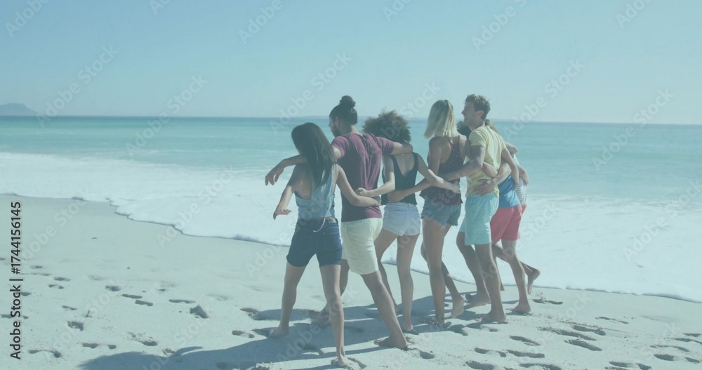 Obraz premium Linking arms seven friends wearing casual attire walking along beach shore, with footprints in sand